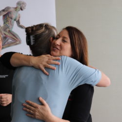 Two people hugging while another person smiles in background.