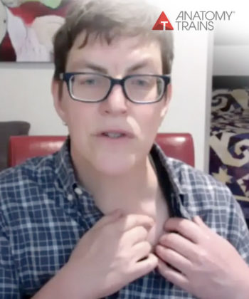 Kieran Schumaker wearing glasses and a plaid button-down shirt looking into the camera and pointing with her index finger to a spot on the upper-right part of her chest.